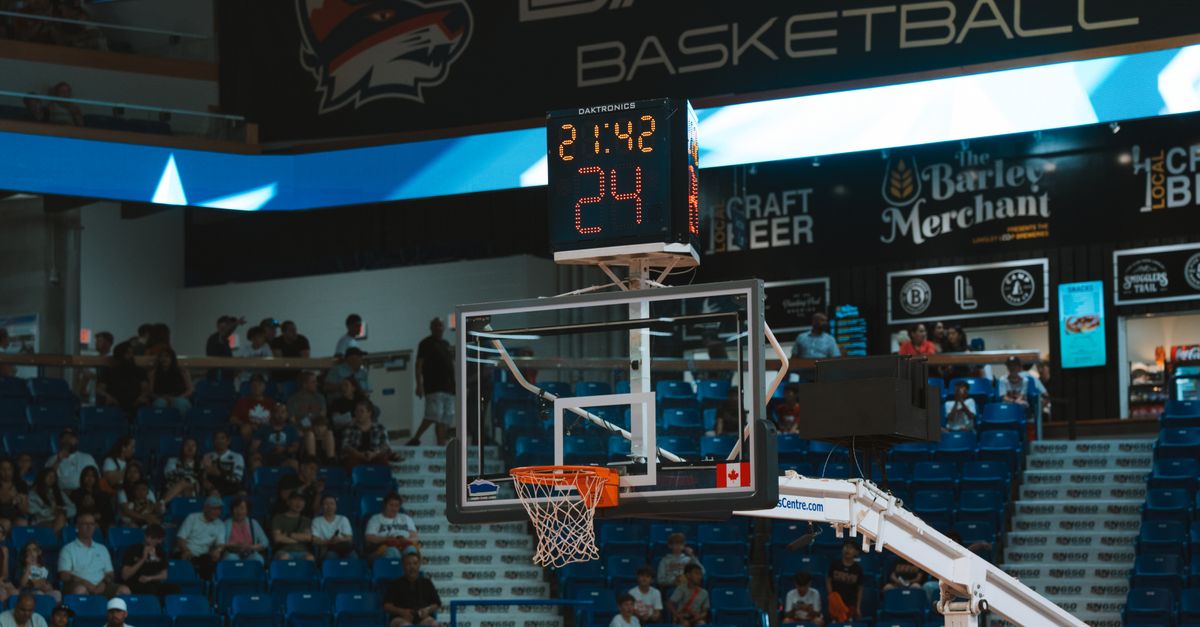 Close-up of a basketball hoop inside a stadium with a scoreboard and audience in the background.